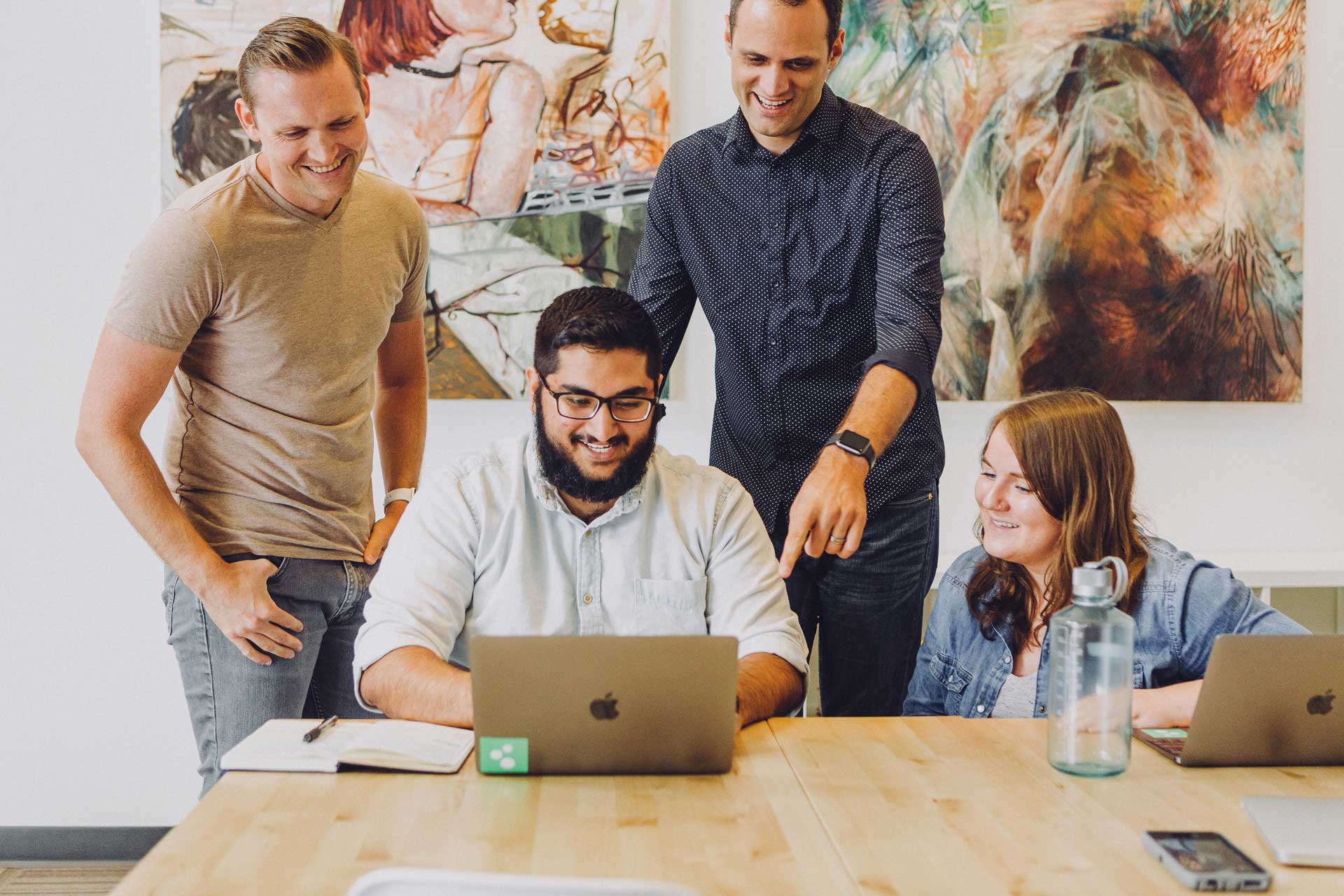 Four cheerful individuals in an office setting, gathered around a laptop.